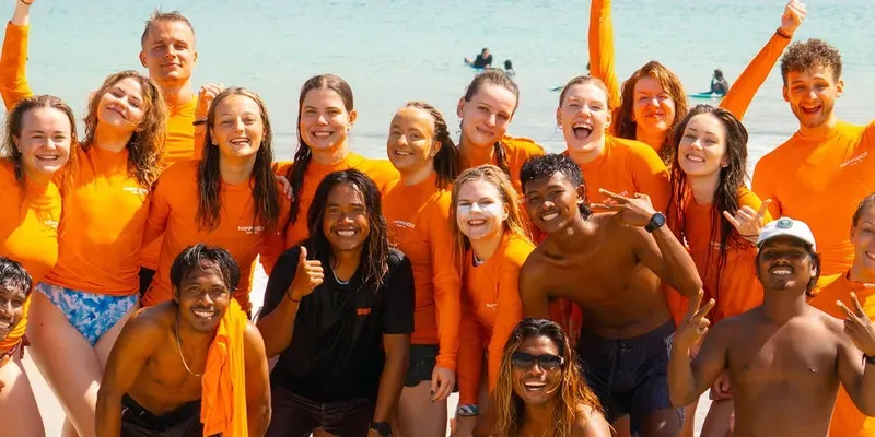The full Happiness Surf Co. crew of coaches and students in orange rashguards cheering together on the beach after a session in Kuta Lombok