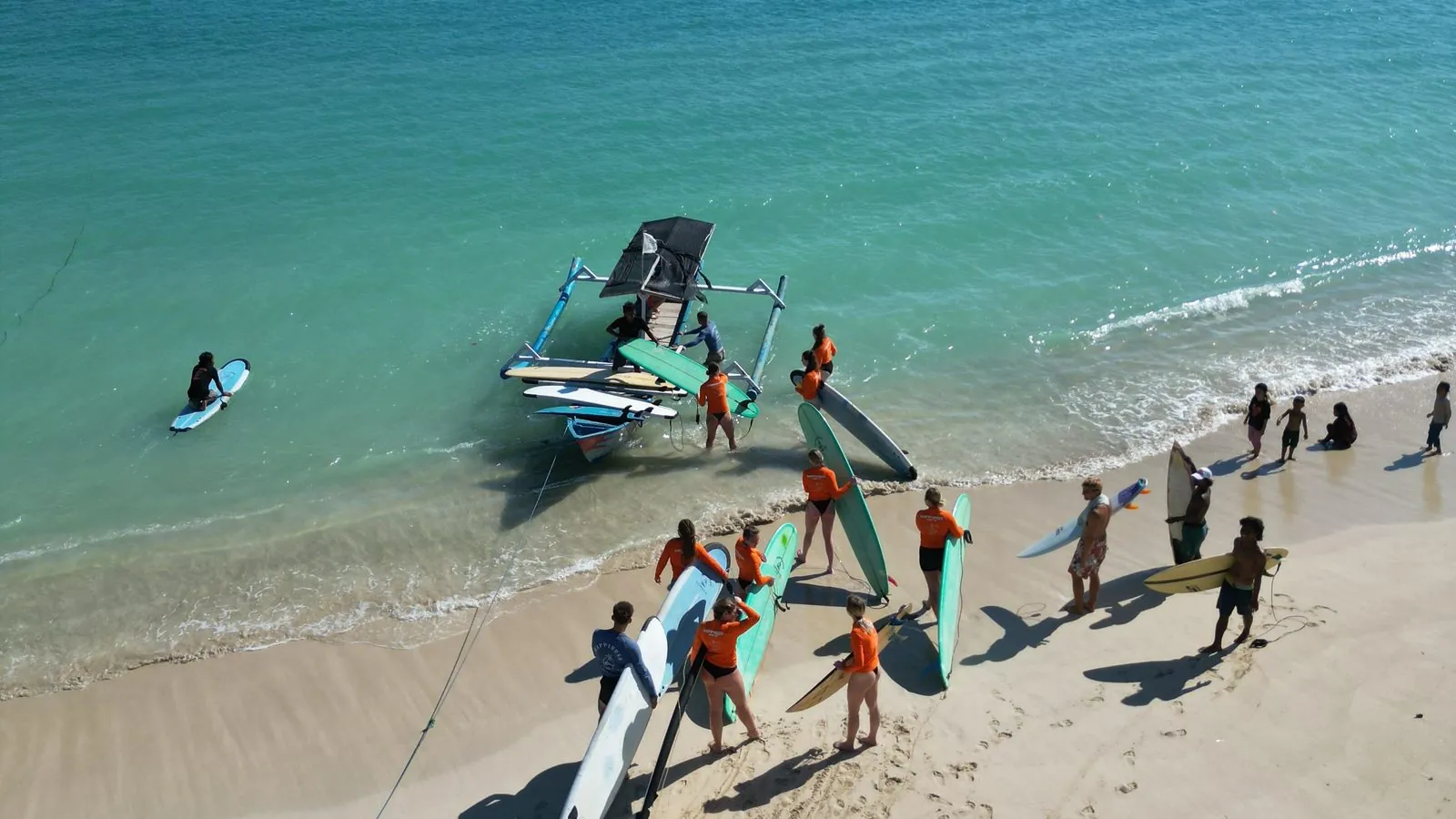 Students in orange Happiness rashguards loading surfboards onto a traditional outrigger boat on a turquoise bay in Lombok