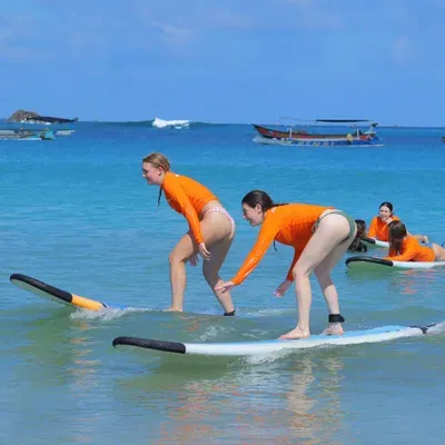 Happiness Surf Co coach guiding a student in an orange rashguard to her feet on a soft-top board in Kuta Lombok