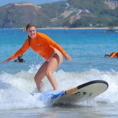 Surf student in an orange Happiness rashguard standing up and riding her first wave in Kuta Lombok
