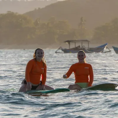 Two surf students in orange Happiness rashguards sitting on their boards in golden-hour light beside traditional boats in Kuta Lombok