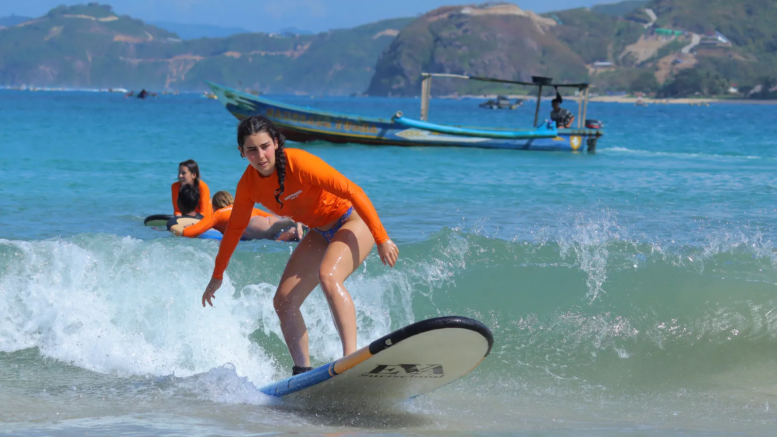 Surf student in an orange Happiness rashguard riding a wave in Kuta Lombok with traditional boats behind her